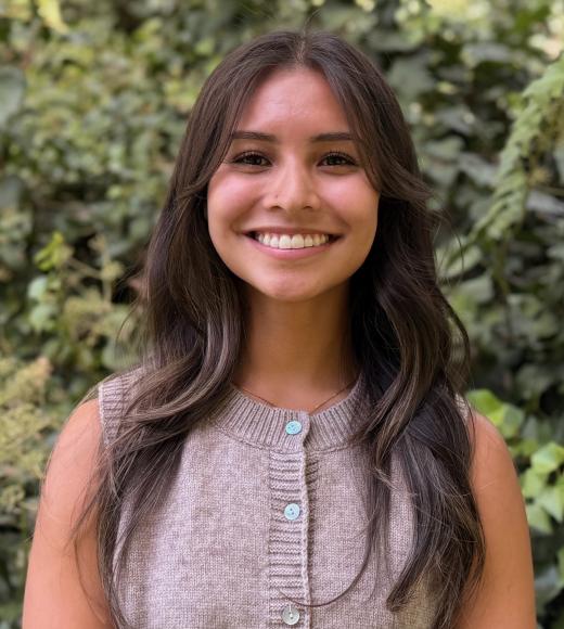 Smiling young woman with long dark hair in sleeveless beige top against leafy green background